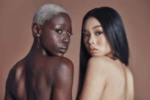 Two young women of African and Asian descent posing back-to-back, showcasing diverse skin tones and high-fashion aesthetics against a neutral background.