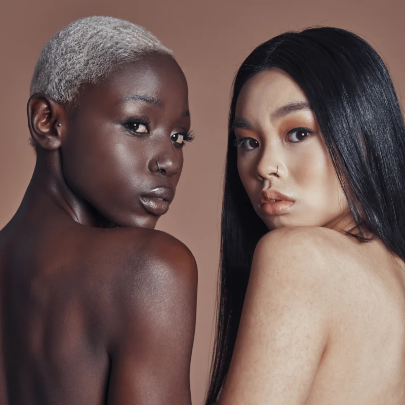 Two young women of African and Asian descent posing back-to-back, showcasing diverse skin tones and high-fashion aesthetics against a neutral background.