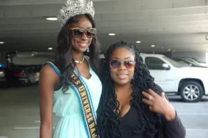 Miss Universe Bahamas 2012 Celeste Marshall, wearing her official sash and crown with sunglasses, standing beside Africa Allah of DIRadioCast/JEM Media Group in a parking structure. This photo documents the arrival and production behind the 25th Annual Atlanta Carnival and Bahamas 40th Independence celebration.