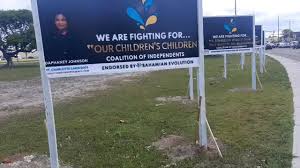 A row of political campaign signs along a grassy roadside in the Bahamas. The signs feature a dark background with a candidate's photo and multiple lines of text, including the slogan "WE ARE FIGHTING FOR... OUR CHILDREN'S CHILDREN" and various organizational endorsements, illustrating the challenge of reading dense text from a moving vehicle.